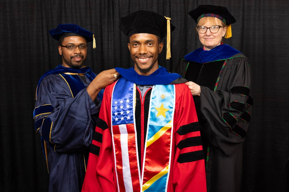 A smiling graduate in a red doctoral gown with stoles featuring the American and Congolese flags is being hooded by two faculty members in academic regalia, all standing in front of a black curtain backdrop.